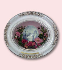 white oval frame with red and pink preserved funeral flowers in arc around old black and white photo of parents with baby
